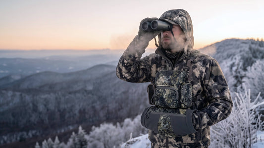 Hunter in camo gear with binocular harness observing snowy mountain terrain at dawn