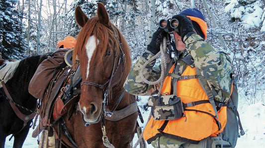 Hunter with binoculars scouting in snowy backcountry, beside a packed horse. Ideal for promoting hunting optics, bino harnesses, and winter outdoor gear.
