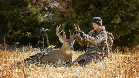 Camouflaged bow hunter kneels beside harvested large whitetail buck in dry farm field, archery big‑game hunting scene with hunting backpack
