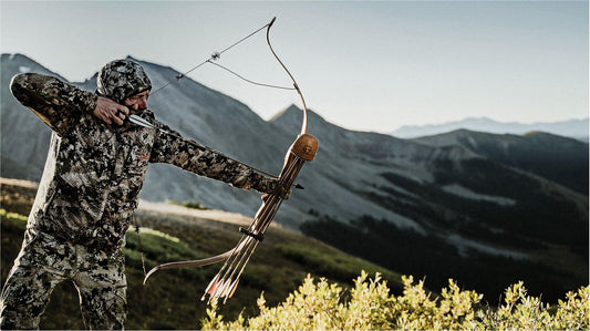 Camouflage bow hunter drawing recurve bow in mountain backcountry terrain for big game hunting