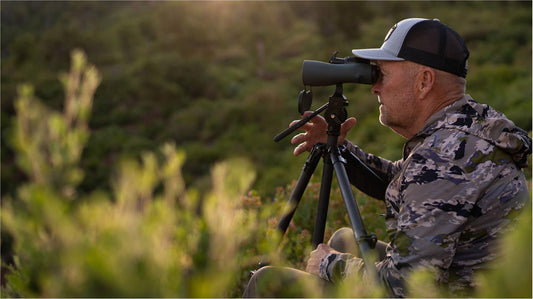 Senior deer hunter in camo uses 10x50 binoculars on tripod for July US hunting, best bino chest pack binoculars harness field scouting scene