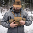 Hunter wearing Moxule coyote brown modular binocular chest harness in snowy woodland, adjusting bino pack, cold weather deer and elk hunting optics gear